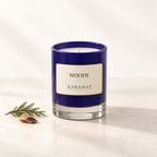 Bougie parfumée Woody, pot bleu et touches discrètes de bois et de feuillage pour évoquer ses notes boisées.